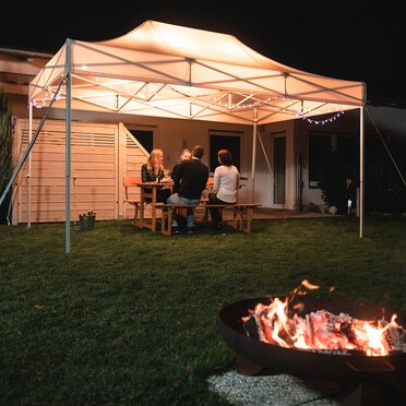 The 4.5x3 m garden tent stands in the garden in the dark. The 4 guests sit under it. There is a fireplace in front of the gazebo.